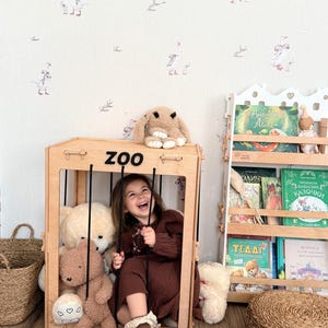 Op de afbeelding: Een jong meisje zit in een houten speelstructuur met het label "ZOO", omringd door knuffels. Een pluchen konijn zit bovenop de structuur. De achtergrond heeft een grillig behang met zwanenillustraties en een boekenplank vol boeken.
