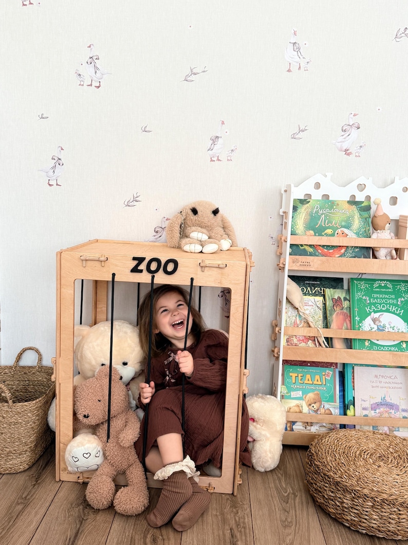 Op de afbeelding: Een jong meisje zit in een houten speelstructuur met het label "ZOO", omringd door knuffels. Een pluchen konijn zit bovenop de structuur. De achtergrond heeft een grillig behang met zwanenillustraties en een boekenplank vol boeken.