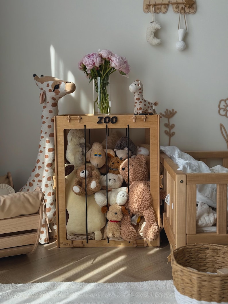 Op de afbeelding: Een houten speelgoedopbergdoos met het opschrift "ZOO" is gevuld met diverse knuffels. Een grote giraf en een kleinere giraf staan links. Een vaas met roze bloemen staat op de doos. Een houten wieg en mand zijn ook in de kamer.