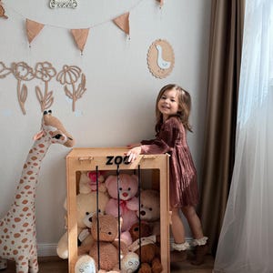 Op de afbeelding: Een houten speelgoed opbergdoos met het label "ZOO" gevuld met teddyberen. Een lange giraf knuffel staat links. Een jong meisje in een bruine jurk staat naast de doos. De kamer is versierd met wandkunst en een slinger.