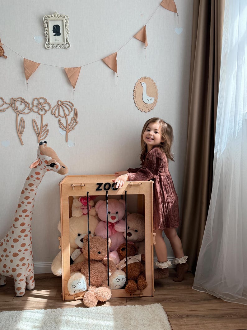 Op de afbeelding: Een houten speelgoed opbergdoos met het label "ZOO" gevuld met teddyberen. Een lange giraf knuffel staat links. Een jong meisje in een bruine jurk staat naast de doos. De kamer is versierd met wandkunst en een slinger.