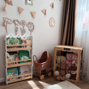 Op de afbeelding: Een kinderkamer met een witte boekenplank vol boeken en knuffels. Een houten speelgoedwandelwagen en een houten speelgoedkist met het woord "ZOO" zijn ook aanwezig. De kamer is versierd met houten bloemenuitsparingen en een banner.