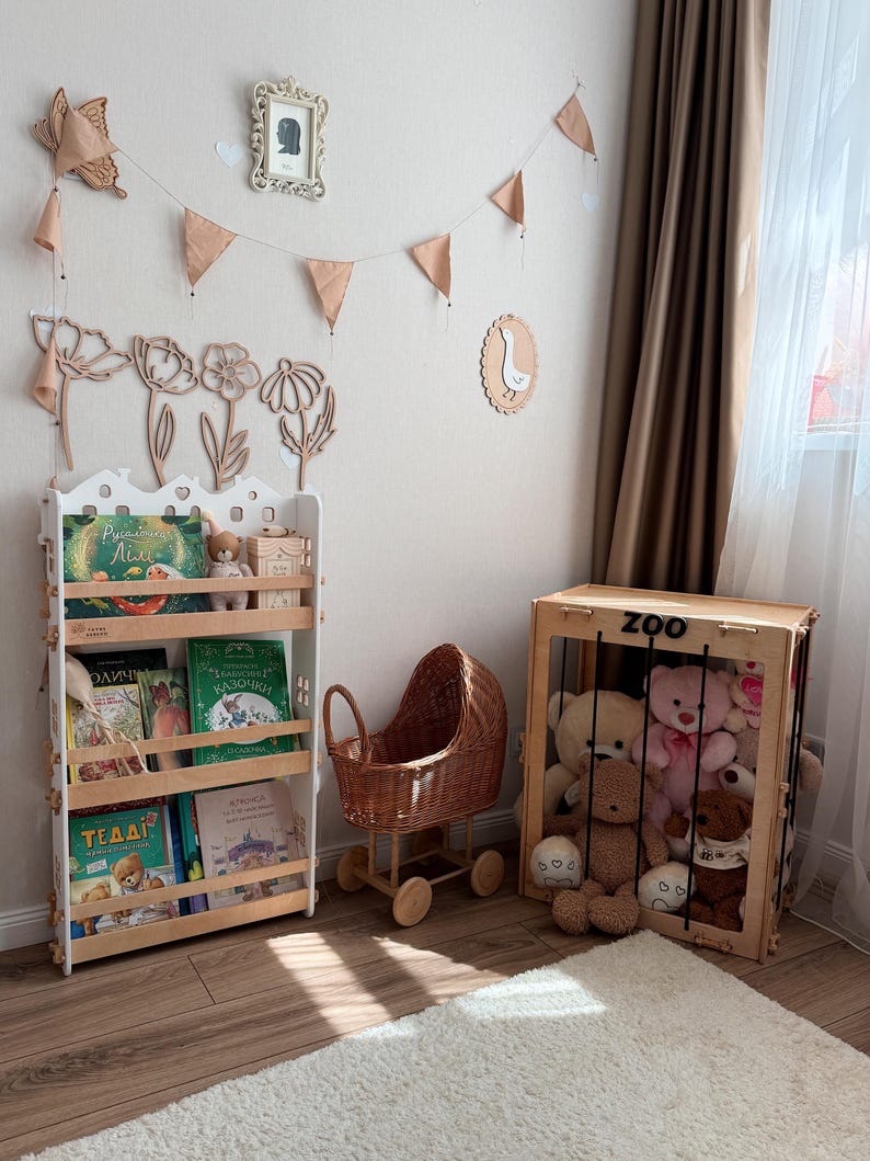 Op de afbeelding: Een kinderkamer met een witte boekenplank vol boeken en knuffels. Een houten speelgoedwandelwagen en een houten speelgoedkist met het woord "ZOO" zijn ook aanwezig. De kamer is versierd met houten bloemenuitsparingen en een banner.