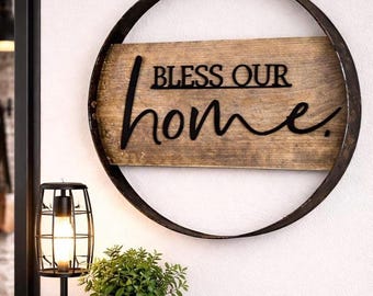 May include: A circular wall sign featuring a dark metal hoop frame surrounding a rectangular wooden plank. The wood has a natural grain finish and displays the black metal lettering that reads BLESS OUR home in a mix of block and script fonts.