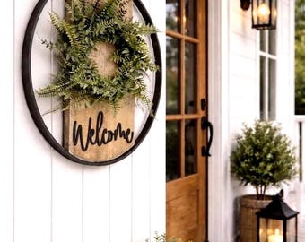 May include: A circular wall hanging featuring a black metal rim enclosing a vertical wooden plank. A green fern wreath is attached to the top half of the wood, and the word Welcome is written in black cursive script on the bottom half.
