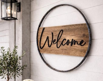 May include: A circular wall sign featuring a dark metal hoop frame surrounding a rectangular piece of natural brown wood. The word Welcome is written across the center of the wood in black, raised, cursive lettering with a smooth finish.