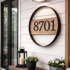 May include: A wooden address sign with the number 8701 in black, set within a dark metal ring. The sign is mounted on a white, horizontal-planked wall, with a black light fixture above. A wooden bench holds a lantern and potted plants.