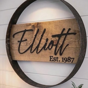 May include: A circular wall sign featuring a dark metal hoop frame enclosing a rectangular wooden board. The board displays the name Elliott in large black script lettering, with the text Est. 1987 printed in a smaller black sans-serif font below.