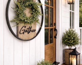 May include: A circular wall hanging featuring a black metal hoop frame surrounding a vertical wooden plank. A green fern wreath is attached to the top half of the wood, and the word Gather is written in black cursive script on the lower half.