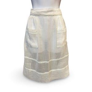 May include: A white apron with two front pockets and lace trim. The apron has green stitching along the edges and pockets. The apron is displayed on a mannequin.