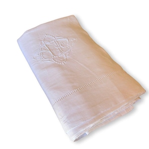 May include: Folded white linen sheet with an embroidered monogram in the upper corner. The sheet has a decorative stitched border along the edge. The fabric appears soft and slightly textured.