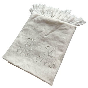 May include: Off-white rectangular scarf with a fringed edge. The scarf features embroidered cursive initials "B M" in the center. The scarf is made of a soft, woven material.