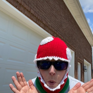 May include: A person wearing a red and white crocheted hat with a green crocheted neck warmer. The hat is shaped like a mushroom and has a white rim. The person is wearing sunglasses and has a surprised expression on their face.