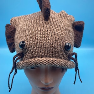 May include: A brown and tan knitted hat shaped like a fish with brown felt fins and black button eyes. The hat has brown yarn dangling from the sides.