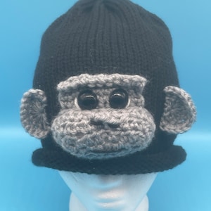 May include: A black knitted hat with a gray crocheted monkey face attached to the front. The monkey face has black eyes and a gray nose.