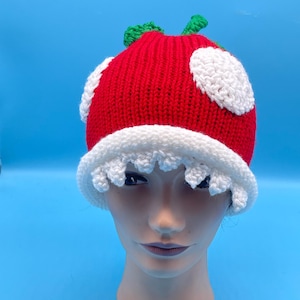 May include: A red and white mushroom-shaped crochet hat with white spots and a white scalloped brim. The hat has green leaves on top and is displayed on a mannequin head against a blue background.