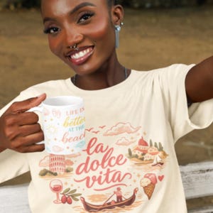 Może przedstawiać: Kremowy t-shirt z kr&oacute;tkim rękawem z nadrukiem graficznym zawierającym napis "la dolce vita" oraz ilustracje gondoli, kieliszka wina, lod&oacute;w w wafelku i włoskiej architektury. W dłoni trzymany jest biały kubek z napisem "LIFE IS better AT THE beach".