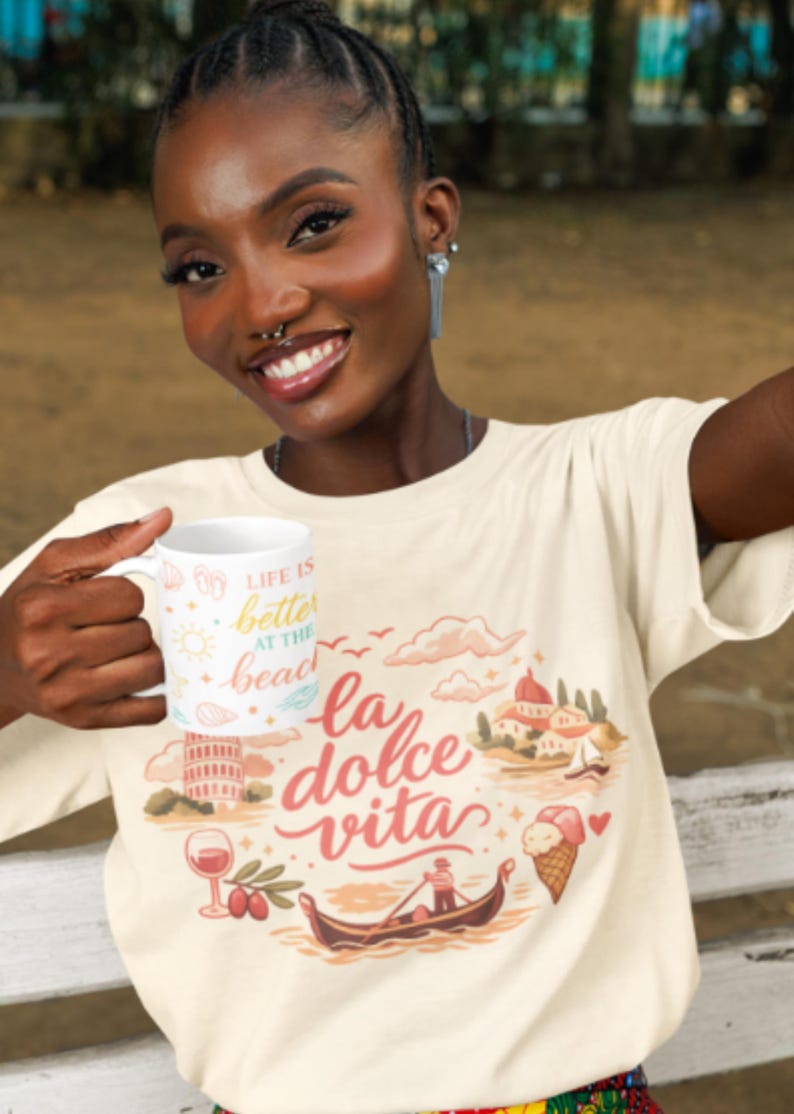 Może przedstawiać: Kremowy t-shirt z kr&oacute;tkim rękawem z nadrukiem graficznym zawierającym napis "la dolce vita" oraz ilustracje gondoli, kieliszka wina, lod&oacute;w w wafelku i włoskiej architektury. W dłoni trzymany jest biały kubek z napisem "LIFE IS better AT THE beach".