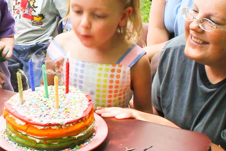 Image shows a child blowing out the candles on her birthday cake. The candles are all rolled beeswax birthday candles handmade by Pumpkin Plus Bear.