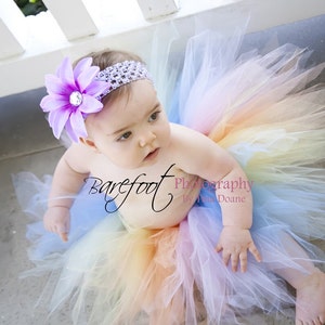 May include: A baby girl wearing a rainbow tulle tutu skirt and a purple flower headband. The skirt is made of layers of tulle in shades of pink, yellow, orange, green, blue, and purple. The headband has a large purple flower with a rhinestone center.