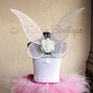 May include: White fairy wings with a white flower accent. The wings are made of sheer fabric and have a delicate design. The wings are attached to a white strapless top and a pink tulle tutu.