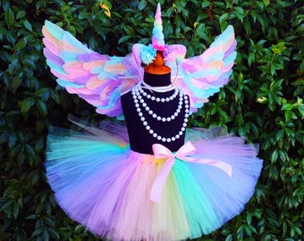May include: A multi-colored unicorn costume displayed on a black mannequin. The costume includes a rainbow tutu, feathered wings, a unicorn horn, and a pearl necklace. The wings and tutu feature pastel colors.