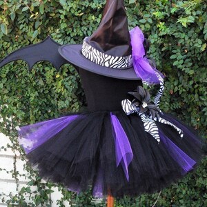 May include: A black and purple tutu with a black witch hat, zebra print ribbon, and bat wings. The hat has a purple bow and a black flower with a rhinestone center.