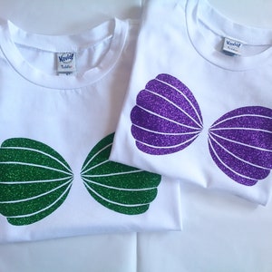 May include: Two white t-shirts with glitter seashell designs. One shirt has green glitter seashells and the other has purple glitter seashells.