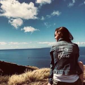 May include: A woman wearing a denim jacket with the text "Mrs. Blom" written in white on the back. She is sitting on a rocky cliff overlooking a body of water with a blue sky and white clouds in the background.