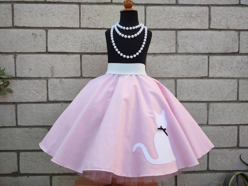 K&ouml;nnte beinhalten: Ein rosa Kreisrock mit einer wei&szlig;en Katzen-Silhouette und einem wei&szlig;en G&uuml;rtel. Der Rock hat einen T&uuml;ll-Unterrock und befindet sich auf einer Schaufensterpuppe mit einer Perlenkette.