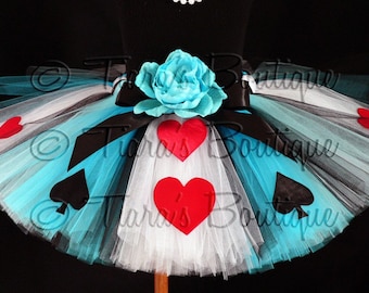 May include: A blue, white, and black tulle tutu with red hearts and black spades. The tutu has a black satin ribbon and a blue flower.