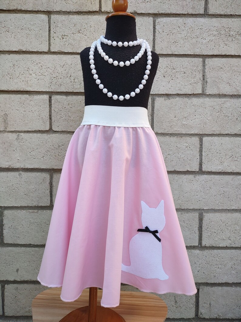 K&ouml;nnte beinhalten: Ein rosa Rundrock mit einer wei&szlig;en Katzen-Silhouette am Saum. Der Rock hat einen wei&szlig;en Bund und befindet sich auf einer Puppe, die eine Perlenkette tr&auml;gt.