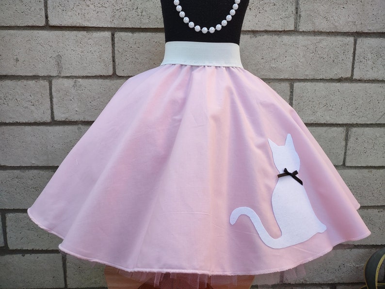 K&ouml;nnte beinhalten: Ein rosa Kreisrock mit einem wei&szlig;en Bund und einer wei&szlig;en Katzen-Silhouette auf der R&uuml;ckseite. Die Katze hat eine schwarze Schleife.