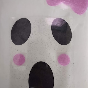 May include: A sheet of stickers featuring a ghost face design with black oval eyes and mouth, and pink glittery cheeks. A pink glitter bow sticker is at the top. The stickers are on a clear background.