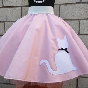 K&ouml;nnte beinhalten: Ein rosa Kreisrock mit einer wei&szlig;en Katzen-Silhouette auf der rechten Seite. Die Katze hat eine schwarze Schleife um den Hals. Der Rock hat einen wei&szlig;en Bund und einen T&uuml;ll-Unterrock.