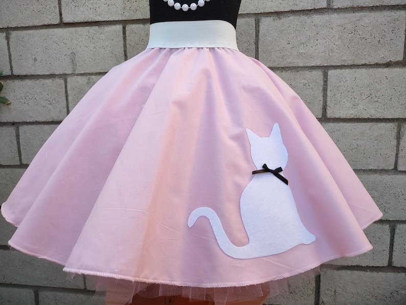 K&ouml;nnte beinhalten: Ein rosa Kreisrock mit einer wei&szlig;en Katzen-Silhouette auf der rechten Seite. Die Katze hat eine schwarze Schleife um den Hals. Der Rock hat einen wei&szlig;en Bund und einen T&uuml;ll-Unterrock.