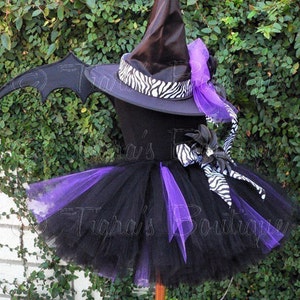 May include: Black and purple tutu with a black witch hat with a zebra print ribbon and a purple bow. The hat has a black bat wing attached to the side.