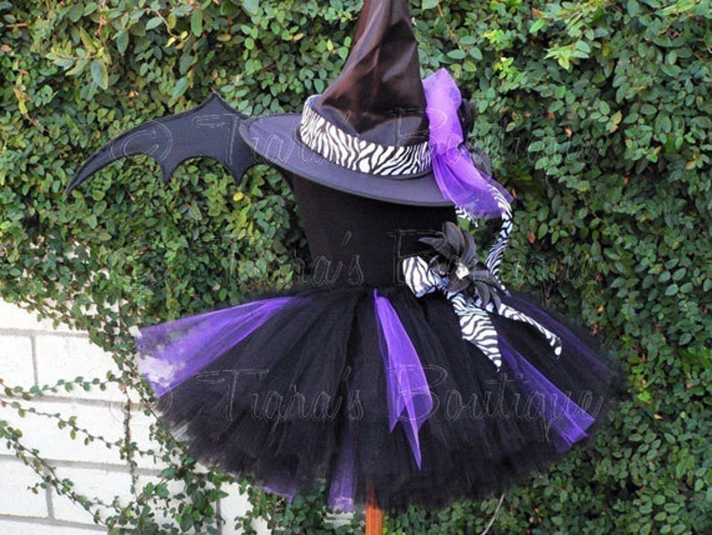 May include: Black and purple tutu with a black witch hat with a zebra print ribbon and a purple bow. The hat has a black bat wing attached to the side.