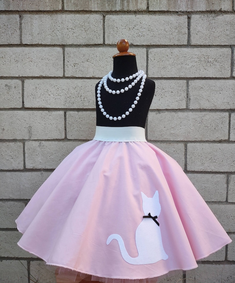 K&ouml;nnte beinhalten: Ein rosa Kreisrock mit einer wei&szlig;en Katzen-Silhouette auf der Vorderseite. Der Rock hat einen wei&szlig;en Bund und wird von einer Puppe mit einer Perlenkette getragen.