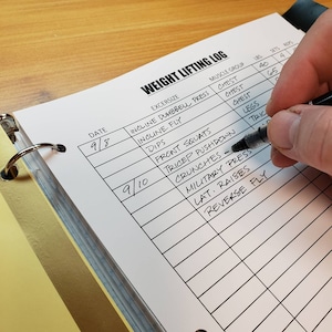 Può includere: Un registro di sollevamento pesi con un raccoglitore nero e una pagina bianca. La pagina ha colonne per la data, l'esercizio, il gruppo muscolare, le libbre, le serie e le ripetizioni. La pagina è piena di voci scritte a mano per diversi esercizi, tra cui panca piana inclinata con manubri, fly inclinato, dips, squat frontali, triceps pushdown, crunch, military press, lat raise e reverse fly.