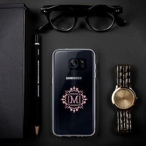 May include: A clear phone case with a pink and green monogram design featuring the letter "M" in a decorative frame. The case is on a black surface with a Samsung phone inside.