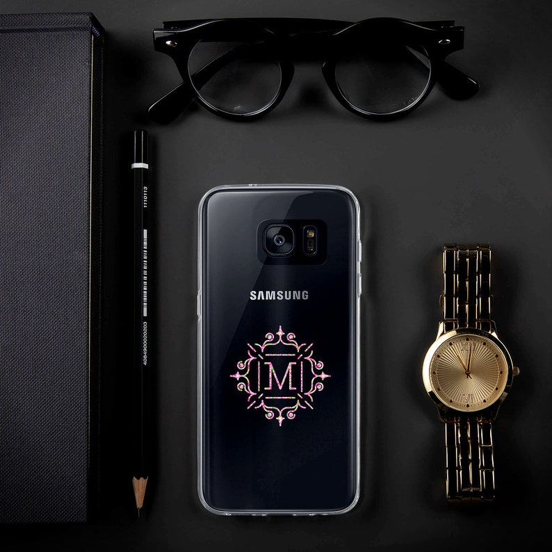 May include: A clear phone case with a pink and green monogram design featuring the letter "M" in a decorative frame. The case is on a black surface with a Samsung phone inside.
