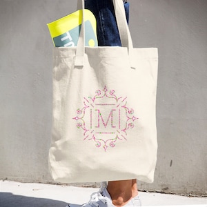 May include: A white canvas tote bag with a pink, green, and white monogram design featuring the letter "M".