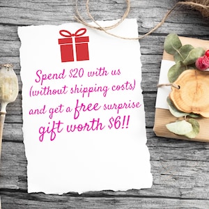 May include: A white paper note with torn edges on a wooden surface. The note has pink text that reads "Spend $20 with us (without shipping costs) and get a free surprise gift worth $6!!". A red gift box icon is above the text.