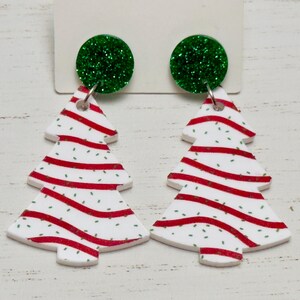 May include: A pair of Christmas tree earrings with red and white candy cane stripes and green glitter accents. The earrings are made of white acrylic and have a green glitter circle at the top.