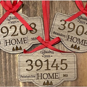 May include: Three wooden Christmas ornaments with a rustic finish. Each ornament features a house-shaped design with a zip code, city, and state. The ornaments are decorated with a red ribbon and a small pine tree design.