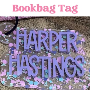 May include: A custom bookbag tag featuring the name Harper Hastings in raised purple lettering. The tag is made of a clear acrylic base filled with purple, light blue, and white hexagonal glitter, and includes a dark purple loop for attachment.