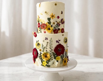 Pode incluir: Um bolo de dois andares com cobertura branca, decorado com flores secas coloridas. O bolo, apresentado em um pedestal branco, apresenta decorações florais vermelhas, amarelas e brancas. Um bolo visualmente atraente para celebrações.
