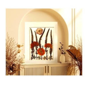 May include: A framed floral arrangement featuring pressed orange and red flowers, with tall, slender brown stems and small blue and white flowers. The white frame is displayed on a white cabinet, with decorative plants in the foreground.