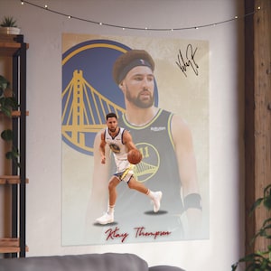 May include: A basketball poster featuring Klay Thompson, a Golden State Warriors player, dribbling the ball. The poster has a yellow and blue background with a silhouette of the Golden Gate Bridge. The poster is signed "Klay" in black ink.
